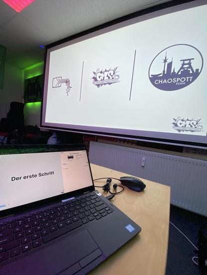Ein Laptop zeigt den Text "Der erste Schritt" auf seinem Bildschirm an, während ein Projektor drei Logos des CCC an einer Wand zeigt.