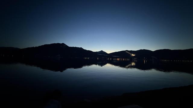 A serene twilight scene over a calm lake, with mountains silhouetted against the deep blue sky. Soft reflections shimmer on the water's surface, illuminated by distant lights from the shore.