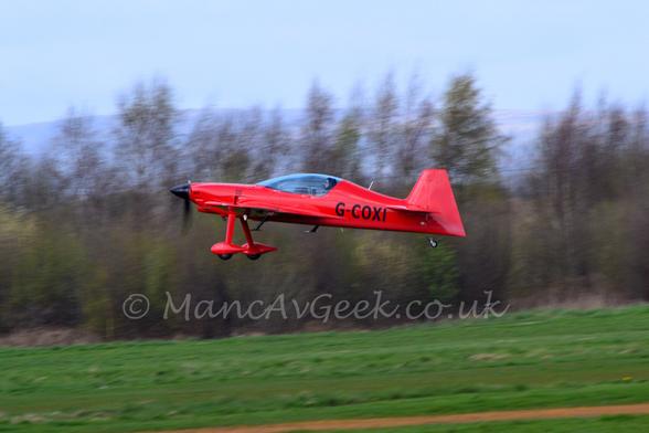 Side view of a small, sleek, single engined light aircraft flying from right to left t a very low altitude, just a few metres above a grass runway.
The plane is a bright scarlet red, with a large glazed canopy over the 2-seat cockpit, with only the rear seat filled.
Long, spindly undercarriage legs extend down from the front of the wings, with large red spats over the main wheels.
The spinner on the propellor is a gloss black, , as is the registration "G-COXI" on the sides of the rear fuselage.
Greengrass fills the foreground at the bottom of the frame, with trees filling the background, with rolling hills and pale blue sky in the distance.