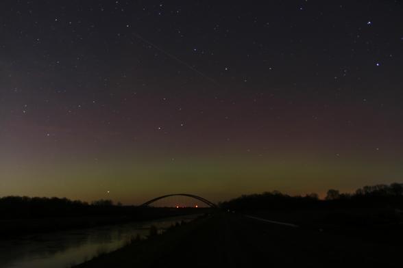 Das Bild zeigt eine Nachtlandschaft mit einem klaren Sternenhimmel. Im Vordergrund befindet sich ein Gewässer, vermutlich ein Fluss oder Kanal, neben dem ein Pfad oder eine Straße verläuft. Am Horizont ist eine Brücke erkennbar, die sich über das Gewässer spannt. Darüber erstreckt sich ein Nachthimmel, der mit zahlreichen Sternen übersät ist. Ein Hauch von grünlichem Polarlicht oder atmosphärischem Leuchten ist ebenfalls sichtbar. Eine helle Linie, möglicherweise eine Sternschnuppe oder Satellitenspur, zieht sich quer über den Himmel. Die Dunkelheit dominiert das Bild, wobei die wenigen Lichtquellen die Szene dezent beleuchten.