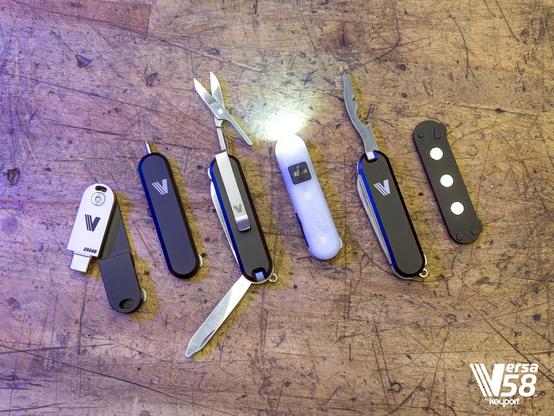 A collection of Keyport Versa 58 modular EDC tools spread across a wooden workbench. The set includes a 256GB USB drive, a multi-tool with scissors and a file extended, a glowing LED flashlight module, and several black modular shells under purple-tinted lighting.