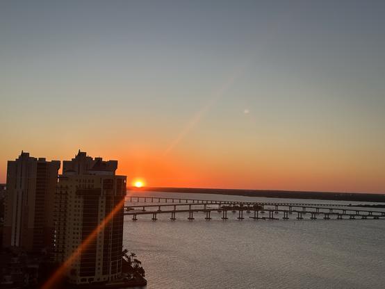 Sun is setting on a black strip of horizon with a wide river. Two taller buildings to the left and a beam of light that cuts from the sun across the buildings to the left and up for the sky on the right. The sky is ombré orange to yellow to blue. Photo by Sewzinski