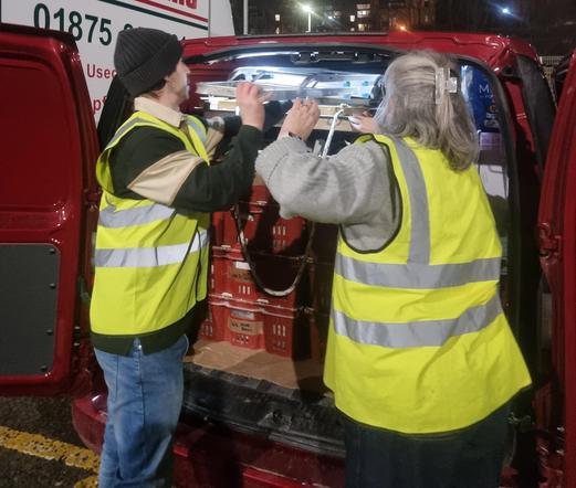 Tracey and Sean, in hi-viz tabards, stand at the back of a red van, lifting a flatbed trolley over the top of stacked red trays of pottery.  It's the last thing to go in the van