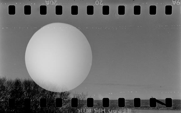 A black and white photo of an outdoor scene with some trees in the left corner and a river bank in the distance to the right and bottom. The most promiment feature is a bright circle almost looks like a giant moon especially since there are many small white dots streaking acoss the frame. The border of the image on top and bottom is perforations from 35 mm film and some film markings. *** Shot with a Mamiya SixVa and Zuiko75f3.5 lens, on IlfordHP5+, and developed with Rodinal by Shom Bandopadhaya. Licensed under Creative Commons Attribution-NonCommercial-ShareAlike (CC BY-NC-SA).