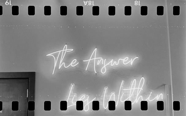 Black and white photo of a neon sign on the wall saying "The Answer Lies Within" with a cursive font. The border of the image on top and bottom is perforations from 35 mm film and some film markings. *** Shot with a Mamiya SixVa and Zuiko75f3.5 lens, on IlfordHP5+, and developed with Rodinal by Shom Bandopadhaya. Licensed under Creative Commons Attribution-NonCommercial-ShareAlike (CC BY-NC-SA).