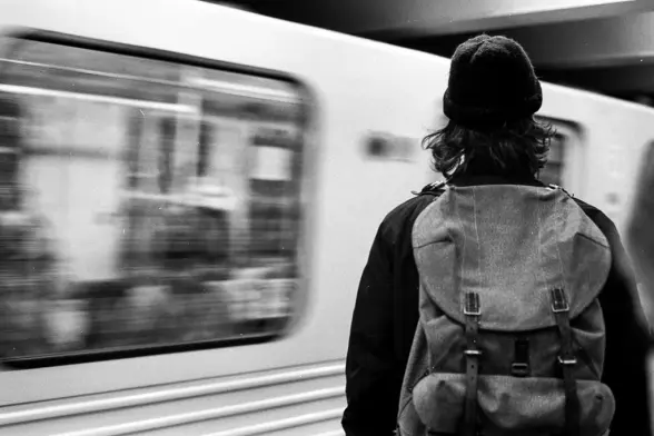 black and white picture with subway wooshing in background and one person seen from behind: long hair, wool hat and an old fashioned backpack.