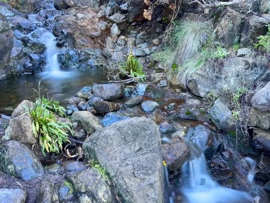 This image shows two small waterfalls using a longer exposure so they are blurred and soft-looking There is a larger upper waterfall that comes down the mountain into a pool, then continues to a smaller waterfall just below. There are a lot of rocks of all sized and a few green plants that have taken root. It's in the shadow of the surrounding hills this time of year. Old Prospector's Trail in the Irish Hills of San Luis Obispo, California.