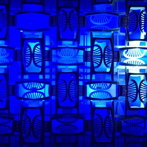 Systematisch ineinander gestapelte Plastikgartenstühle. Es ergibt ein Muster aus Rechtecken, muschelartigen Elementen aus den Rückenlehnen und Verbindungsstücken. Das Werk ist blau beleuchtet.