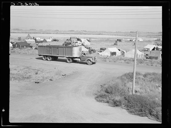 The image is a black and white photograph depicting an outdoor scene with numerous makeshift tents or shacks, likely part of a migrant worker camp. There's one prominent truck parked on the left side of what appears to be a dirt road leading through the area. The trucks are older models suggesting this could be historical photo from past decades. No people can be discerned in these structures which indicates it may not capture daily life within but instead an external view, perhaps taken at night or during non-busy hours due to lack of visible activity around them.
The terrain is relatively flat with sparse vegetation and no signs indicating proximity to water sources such as rivers or lakes. The image has a timestamp overlay in the top-left corner which appears to read "8-2305," possibly denoting August 23, 1947 based on standard format for dates at that time period.
In terms of context given by the caption and title provided, this might be part of a larger series documenting conditions faced during wartime or post-war migration periods when agricultural laborers moved in large numbers to harvest crops. The setting could likely correspond with regions known for agriculture such as California's Central Valley which is often associated with potato picking camps like referenced here.
The visual composition seems straightforward without any dramatic angles, focusing on the s [...]