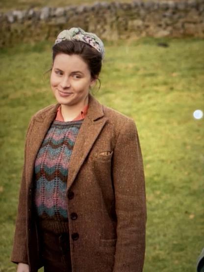 A shrewdly smiling young woman attired wonderfully in rustic richly colored Yorkshire woolens, alone in a green pasture