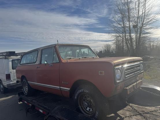 An orange vintage SUV is parked on a trailer, partially surrounded by other vehicles. The rear of the SUV features a large window, and it has a slightly weathered appearance. The background includes trees and a cloudy sky.