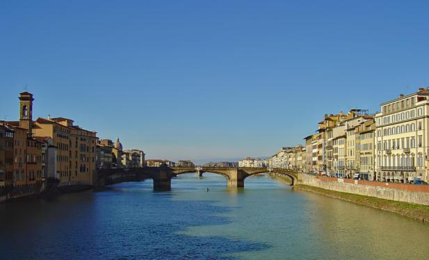 Blue sky over a river flowing through a city with historical multicolored buildings lining both sides. A stone bridge arches across the river in the distance. The city's architecture includes visible features such as shuttered windows and ornate facades.