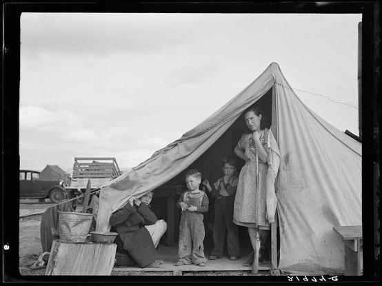 The image shows a family of four individuals inside and near a makeshift tent. The structure is simple, with fabric walls supported by wooden poles and stakes into the ground for stability.

Within the tent, there's a woman in front who appears to be standing guard or observing their surroundings attentively. She has her hand raised up close to her face, possibly wiping her nose or mouth. Her expression seems contemplative or worried. The other three individuals are outside of the tent: one child is seated on wooden boxes with his head down and eyes closed as if he might be asleep or resting; another small boy stands beside him looking out towards something beyond the frame with a neutral posture, while an older girl holds what looks like a broom upright in her right hand. They all wear long-sleeved shirts suitable for cooler weather.

Outside of the tent is another adult man seated on wooden boxes pouring contents from one bucket into another. He's dressed warmly and seems engaged in his task with focused attention to it, indicating that he might be trying to cook or prepare a meal despite their circumstances. In front of him lies an old-fashioned metal bucket filled with what could possibly be food supplies.

The environment around them appears quite barren; there is not much vegetation visible except for some grass patches on the ground and part of another tent in the  [...]