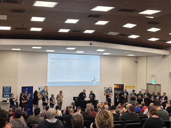 Lots of people standing at the front of a lecture theatre in Everything Open Volunteer shirts, with a slide on the screen listing all their names. The audience is applauding.