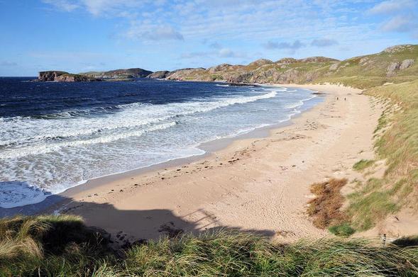 A view of the stunning beach at Oldshoremore. The photo is taken from dunes above the beach and the sand curves round to the left, with waves breaking and a dark blue sea. The headland at the far end is stony. Two tiny figures can be seen walking, some way along the otherwise deserted beach.