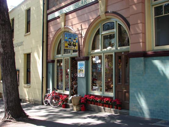 Zwei Bogenfenster eines Ladengeschäfts in einer hellroten Fassade an einem Gehweg. Auf einem Schild zwischen den Bögen oben ist die Zeichnung zweier Bären an einem Strand erkennbar, darüber der Schriftzug "Aussie Bear & Friends", der sich auch an der Fassade wiederfindet. Im Fenster verschiedene Teddybären, auf der Stufe vor den Fenstern eine ganze Reihe von Weihnachtsstern-Pflanzen in Töpfen sowie angelehnt ein rotes Fahrrad. Die Sonne scheint durch Lücken in einer Baumkrone auf die Fassade.