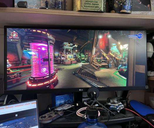 Photo of a wide-screen computer monitor displaying the game Deep Rock Galactic. A small joypad, a water bottle, part of another screen, some things on a shelf (including a tin full of dice, a different tin full of dice, and a jar full of dice), cables, more chewing gum than I expected, and general desk clutter can also be seen. Needs a good dusting, now I look at it…