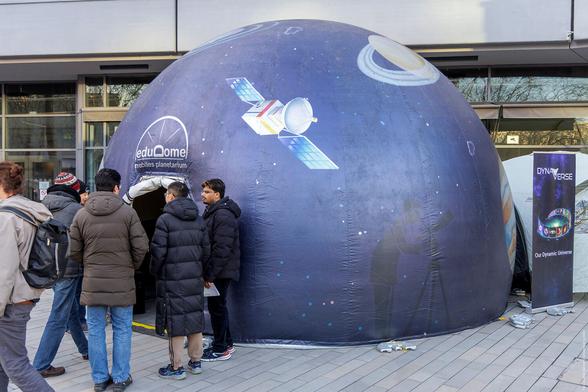 Aufblasbares mobiles Planetarium mit Abbildungen von Satelliten und Planeten. Menschen stehen davor in einer Reihe.