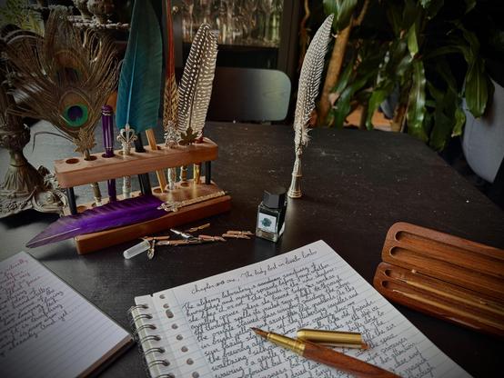 Various funky quills, pens, and paper with cursive handwriting on it on a black table.