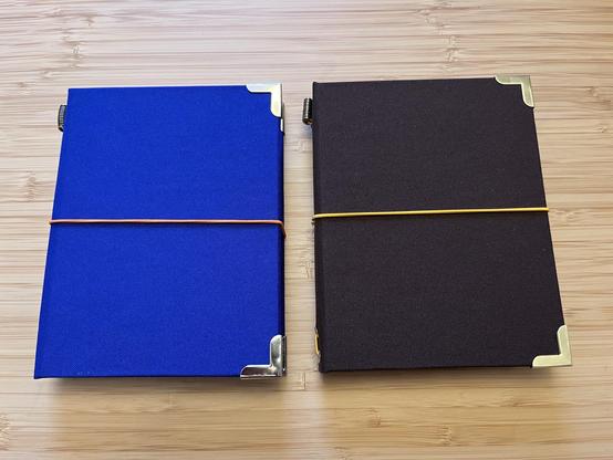 The image features two handmade traveller’s journals placed side by side on a bamboo table. The left notebook is blue with an orange elastic band, and the right notebook is dark brown with a yellow elastic band. Both notebooks have metal corner protectors and both have cloth covers.