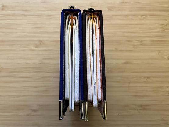 Two traveller’s journals are placed side by side on a bamboo table. The photo is from the top so you can see the way the elastics are woven. Each book contains three passport sized notebooks. The journals are blue and brown and the elastics are orange and goldenrod respectively.