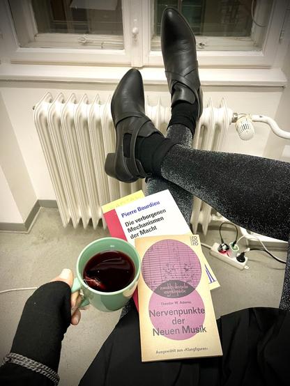This is a somewhat shitpost-ey photograph, so don‘t read too carefully. I sit on a chair wearing a black pleated mini skirt, some stainless steel chain dangling over to where I wish something would‘ve been right from my birth, but I digress. Glitzy silver-lined black tights cover my legs down to black, high heeled ankle boots, the latter resting on the radiator beneath a generic university institute window. On my crossed legs rest two books, one is Theodore W. Adorno‘s „Nervenpunkte der Neuen Musik“, a rate vintage book of 1959, and the other is Pierre Bourdieu‘s „Die verborgenen Mechanismen der Macht“. By now you might‘ve guessed I study and write music sociological theory but anyway, I hold a cup of tea in my left hand, which is covered by a black turned-up cuff, which in turn renders the stainless steel chain there even more prominent. Yes, I‘m somewhat bored and not quite motivated, so enjoy a different kind of text work here …