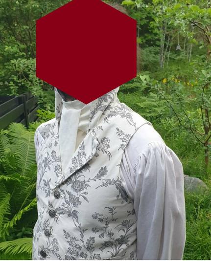 Kukalliseen valkoiseen liiviin pukeutunut mies seisoskelee kesämaisemassa leveähihaisessa paidassa.