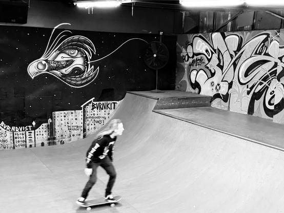 A skateboarder in a black long sleeve with long hair is riding a half-pipe ramp inside a skate park, accompanied by vibrant graffiti on the walls, including a stylized bird design and various colorful tags. The image is in high contrast black and white.