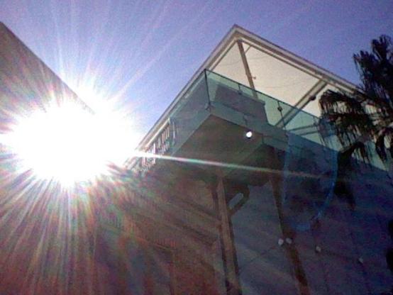 Photo of the sun peaking behind a tall square brick-and-glass building. The sun is directly visible to the camera and blasts the image with brilliant beams of warm-white and rainbows. The flare also created a ghostly blue oval and a small bright dot on the opposite side of the sun.