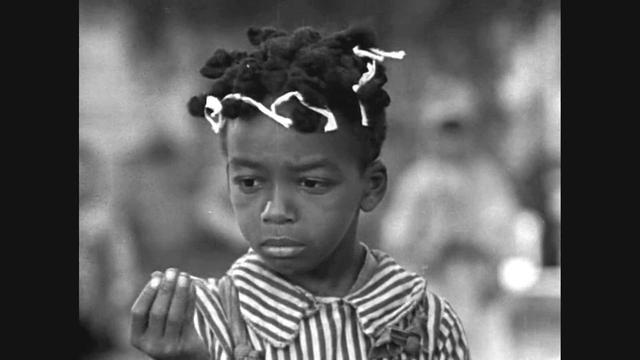 Black-and-white film still of Allen “Farina” Hoskins wearing a striped shirt, looking down at his hand with a serious expression. Blurred figures are visible in the background.