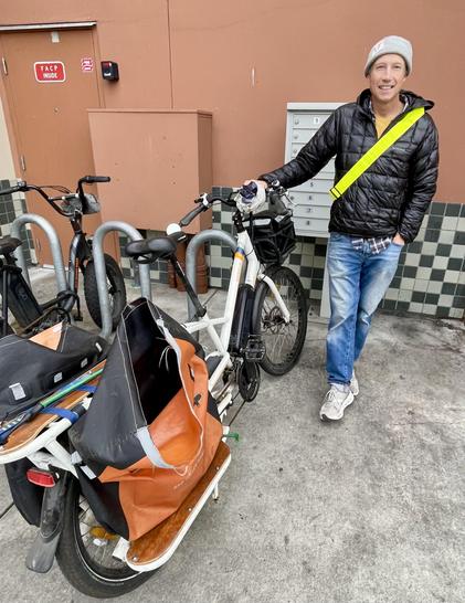 Two bicycles parked on a rack in the "Uptown Monterey" shopping center on chilly afternoon of 23 January 2026. A man stands with his hand on his bicycle, a cargo bike with two orange panniers at rear and and a black basket in front. He is beaming with the joy of biking!