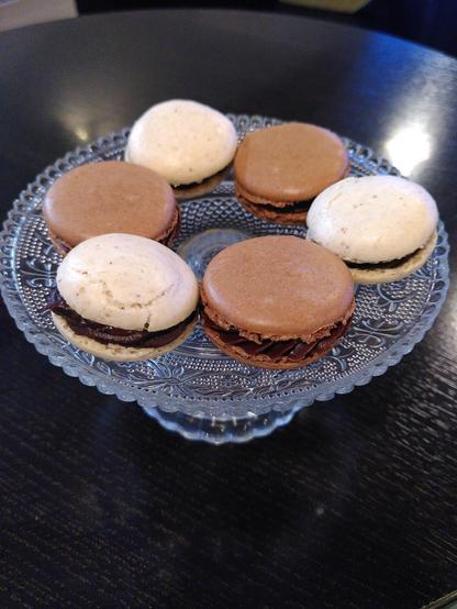 Auf einem dunkelbraunen, runden Holztisch im Café steht eine kleine Glasplatte mit Fuß. Darauf thronen sechs Macarons (feine Makronen aus Mandeln bzw. Nüssen) in zwei Farben (crèmefarben und schokobraun) mit unterschiedlichen Füllungen. Bei den hellen Plätzchen handelt es sich um Makronen mit Haselnüssen, gefüllt mit einer dunklen Schokoladenganache mit Karamell sowie einem Klecks Karamell in der Mitte oder einer Vollmilchschokoladen-Haselnuss-Ganache. Die braunen Macarons sind mit Mandeln und Kakao gebacken und gefüllt mit entweder einer Vollmilchschokoladenganache mit Earl-Grey-Tee oder einer Edelbitterschokoladen-Orangen-Ganache. Die Plätzchen haben außen eine zarte Kruste, innen sind sie weich und etwas klebrig. Die Füllungen sind samtig-weich und wunderbar schokoladig.