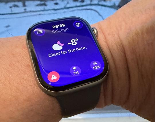 Close-up of a person’s wrist wearing a black smartwatch that displays a weather app showing Chicago at 06:59 with a dark blue screen, clear sky icon, and temperature of −8 degrees against a snowy outdoor background.