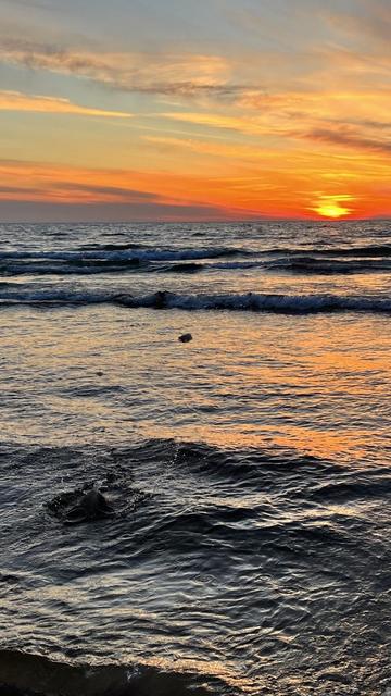 A red and orange sunset over Lake Michigan. The sun is sitting right above the water. In the foreground a flat piece of ice hovers 2 feet over the water and you can see the circular splash where it just skipped off of the water.