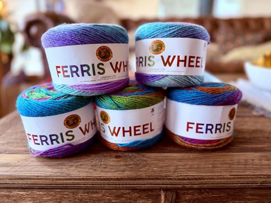 Five skeins of Lion Brand “Ferris Wheel” yarn sit on a wooden surface, each featuring a vibrant multicolor blend.