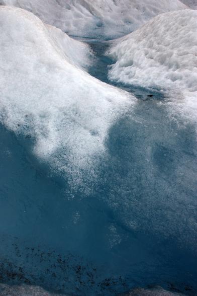 Closeup view of a glacier's crevices in the ice and deep teal water filling those crevices. The ice is white and in mounds, and the water has ice under its surface, smooth teal water as well as black patches of dirt. Juneau, Alaska