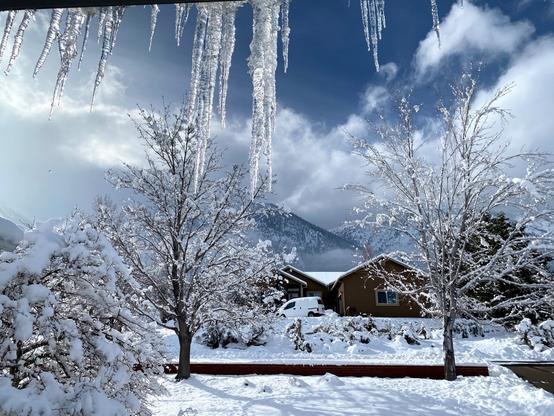 Icicles hang from the eaves of a house at the top of the frame. Beyond, the ground is covered with several inches of snow and chunks of snow and ice cling to the trees. In the distance are snow-covered mountains with abundant white clouds moving over the tops. A few patches of blue sky are also visible. Eastern base of the Sierra Nevada mountains near Lake Tahoe/Carson City, Nevada.