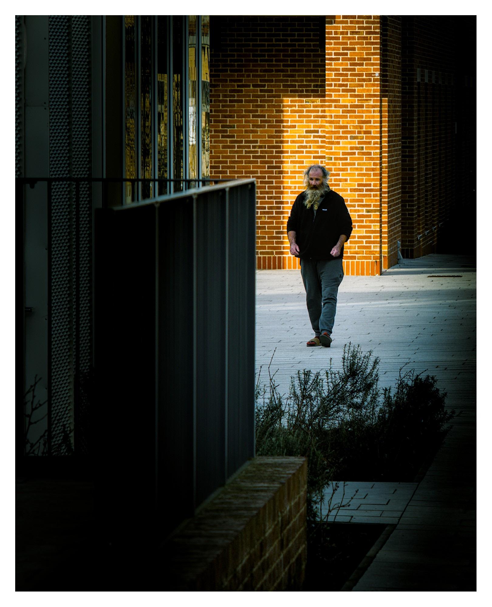 Bearded man walking towards viewer. Light illuminates a brick all behind him. Modern buildings surround him.