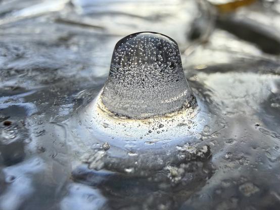 Ein Nahaufnahmebild einer abgerundeten Eisformation mit eingeschlossenen Luftblasen und einer glänzenden Oberfläche, die Licht reflektiert. Der Hintergrund ist verschwommen mit einem Hauch von Farbe unter dem Eis.