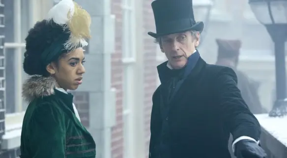This is a photo of Pearl Mackie and Peter Capaldi from Doctor Who. She's in a velvet green dress and he's in a black velvet jacket, top hat, and blue waistcoat. He has a cravat around his throat.