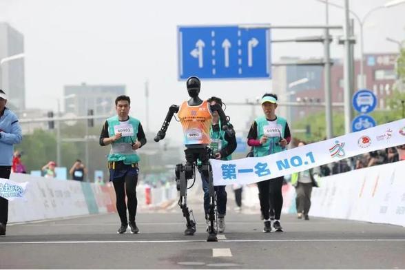 <p><span style="font-weight: 400;">2025年4月、北京市亦庄区で初めて開催された「人型ロボット・ハーフマラソン」が、世界的な注目を集め、人型ロボット企業にとって最高の舞台となった。</span></p>
<p><span style="font-weight: 400;">その第2回大会となる「2026 北京亦庄人型ロボット・ハーフマラソン」の開催が決定した。2026年4月19日午前7時30分の号砲とともにスタートし、同日には人間のランナーが参加する「北京亦庄ハーフマラソン」も併催される。</span></p>
<p><span style="font-weight: 400;">大会は前回に続き「人機共走」方式を採用し、人間の選手と人型ロボット（ヒューマノイド）が同時にスタートし、同一のコースを使用するが、安全確保のため鉄柵や緑地帯によって分けられる。また、海外チームの参加枠を積極的に拡充し、国・地域を問わず参加チームを募集する方針だ。</span></p>
<blockquote class="wp-embedded-content"><p><a href="https://36kr.jp/343456/">世界初「人型ロボット・マラソン」、北京で開催。21km激走レースに20チームが参戦、完走はわずか6体【Update】</a></p></blockquote>
<p></p>
<h3><b>自律制御と遠隔操作を明確に区分</b></h3>
<p><span style="font-weight: 400;">注目すべき点として、初回大会と異なり、2026年大会では自律制御部門と遠隔操作部門が設けられる。同一会場で競技が行われるが、別々に記録が計測されて、より実践的なロボット技術が評価されることになる。</span></p>
<p><span style="font-weight: 400;">初回大会では、多くの参加チームは、人間の操作員がロボットと並走し、リモコンで遠隔操作する方式を採用していた。また、一部では、ロボット選手が操作員の身体に装着された信号装置を「見ながら」、その信号の誘導によって全行程を走破していた。</span></p>
<p><span style="font-weight: 400;">主催側は今回の制度変更を、人型ロボットを「遠隔操作」から「自律制御」へと進化させる重要なステップと位置付けている。</span></p>
<h3><b>技術評価の重点は4分野</b></h3>
<p><b>2026年大会では、技術的ブレークスルーの面で、以下の4つの方向に重点を置く。</b></p>
<h4><b>1.自律性の向上</b></h4>
<p><span style="font-weight: 400;">非自律完走ロボットのチーム成績への点数を調整すると同時に、遠隔操作・ナビゲーション要員が不要な介入を厳格に規制し、ルール面からより多くのロボットが自律完走を実現するよう誘導する。</span></p>
<h4><b>2.航続能力の強化</b></h4>
<p><span style="font-weight: 400;">エネルギー効率の向上に注力し、電池交換の回数を最小限に抑えた設計で、長距離・安定運転の限界に挑戦することを促す。</span></p>
<h4><b>3.擬人性の評価</b></h4>
<p><span style="font-weight: 400;">バイオミメ