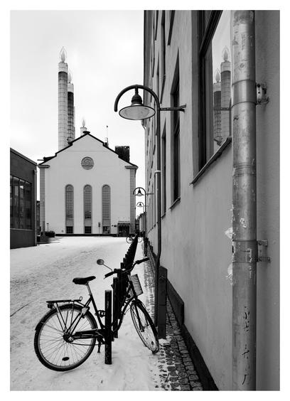 En stående svartvit bild på ett värmeverk som ser ut som en kyrka. Gaveln har 3 avlånga fönster och ett litet runt högre upp. På taket står tre skorstenar med ”ljuslågor” högst upp så de ser ut som stearinljus. I förgrunden en parkerad cykel under en lampa vid ett hus. Snö på marken.