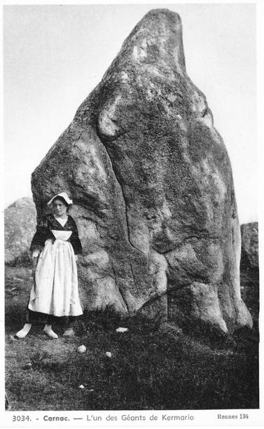A woman in Breton dress stands in front of an enormous megalith with a tapered peak. The caption reads: "3034 - Carnac - L'un des Geants de Kermario    Rennes 134"