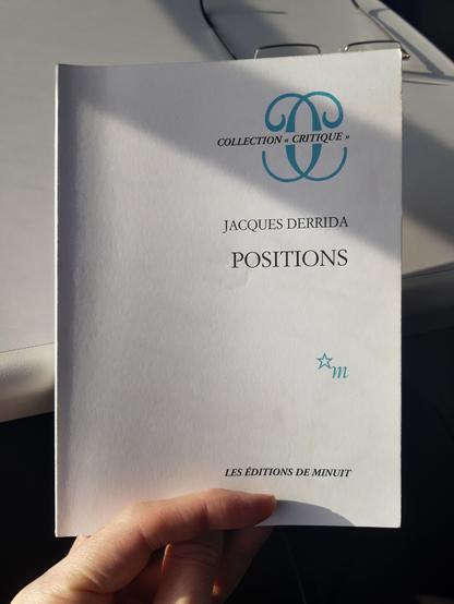 A hand holds a copy of the book "Positions" by Jacques Derrida.
