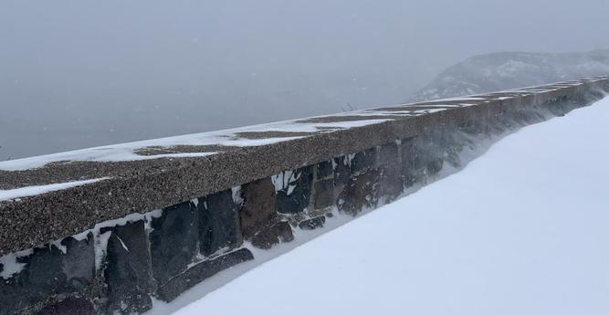 A low stone wall separates a snowdrift-covered sidewalk from a dense grey coastal ocean view. The wall crosses from bottom left to too right and is made of rectangular stones, dark grey and dark red, with a brown red concrete cap. Snow sticks in the crevasses and in patterns on the top. Wind swirls snow from the sidewalk drifts over the wall, with a snow covered hill top barely discernible in the background. [Signal Hill National Historic Site (St. John’s, Canada) January 2026]
