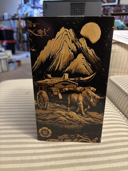 Side of the Transformers Timelines samurai Optimus Prime and Starscream box, showing Optimus in his oxcart mode. The illustration is gold on a black background, with a mountain in the background and the sun behind that.