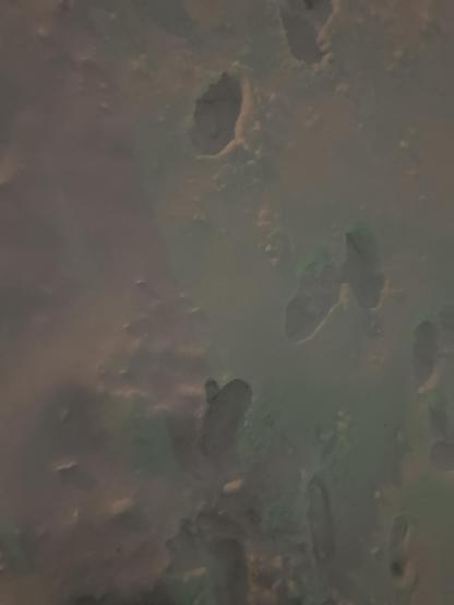 A close-up view of snow-covered ground with several footprints pressed into it. The footprints vary in depth and shape, with one deeper oval-shaped print near the upper middle and a few partial shoe prints toward the right and bottom. The lighting looks dim and slightly yellow-green, like nighttime street lighting reflecting on the snow. No people or other objects are visible.

Image description provided with Be My Eyes.