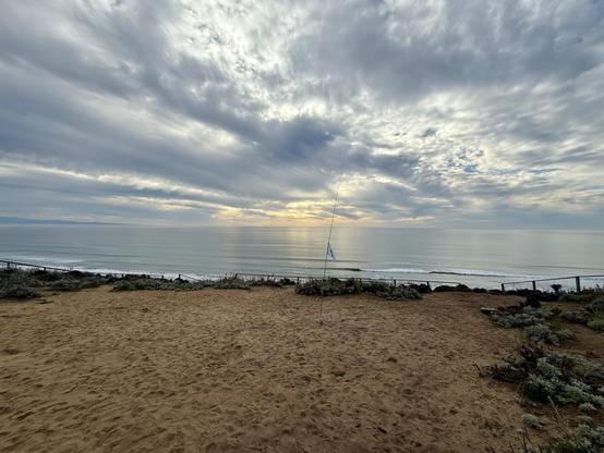 A vertical antenna set up on a sandy area on top of a hill right next to the beach. In the background is the pacific with some waves and a very cloudy sky.