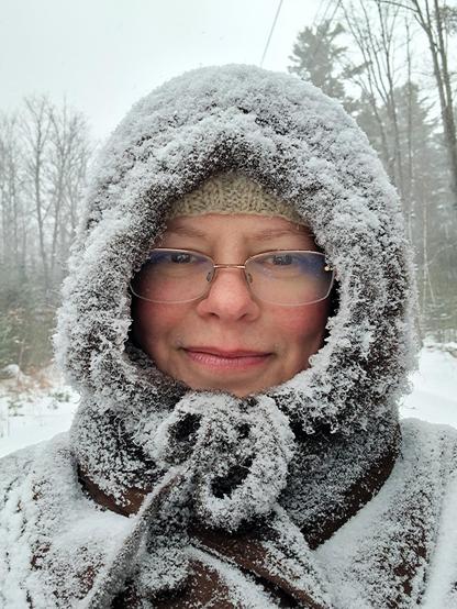 A person (me) in shearling cape, yak-fur snood, and glasses The clothes are covered in snow such that you cannot tell their colors.. My cheeks are pink, my glasses are slightly fogged, and I look pretty happy. Behind me is a rural scene of trees and blowing snow.