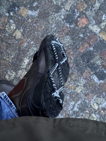A person wearing a brown boot with black laces and a black sole stands on a cobblestone surface.the soles of the boots are covered with snow chains that are strapped on the boots.